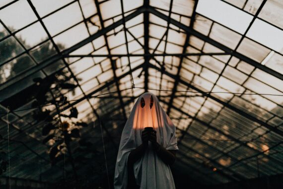 Mysterious figure in ghost costume holding lantern inside a dimly lit greenhouse.